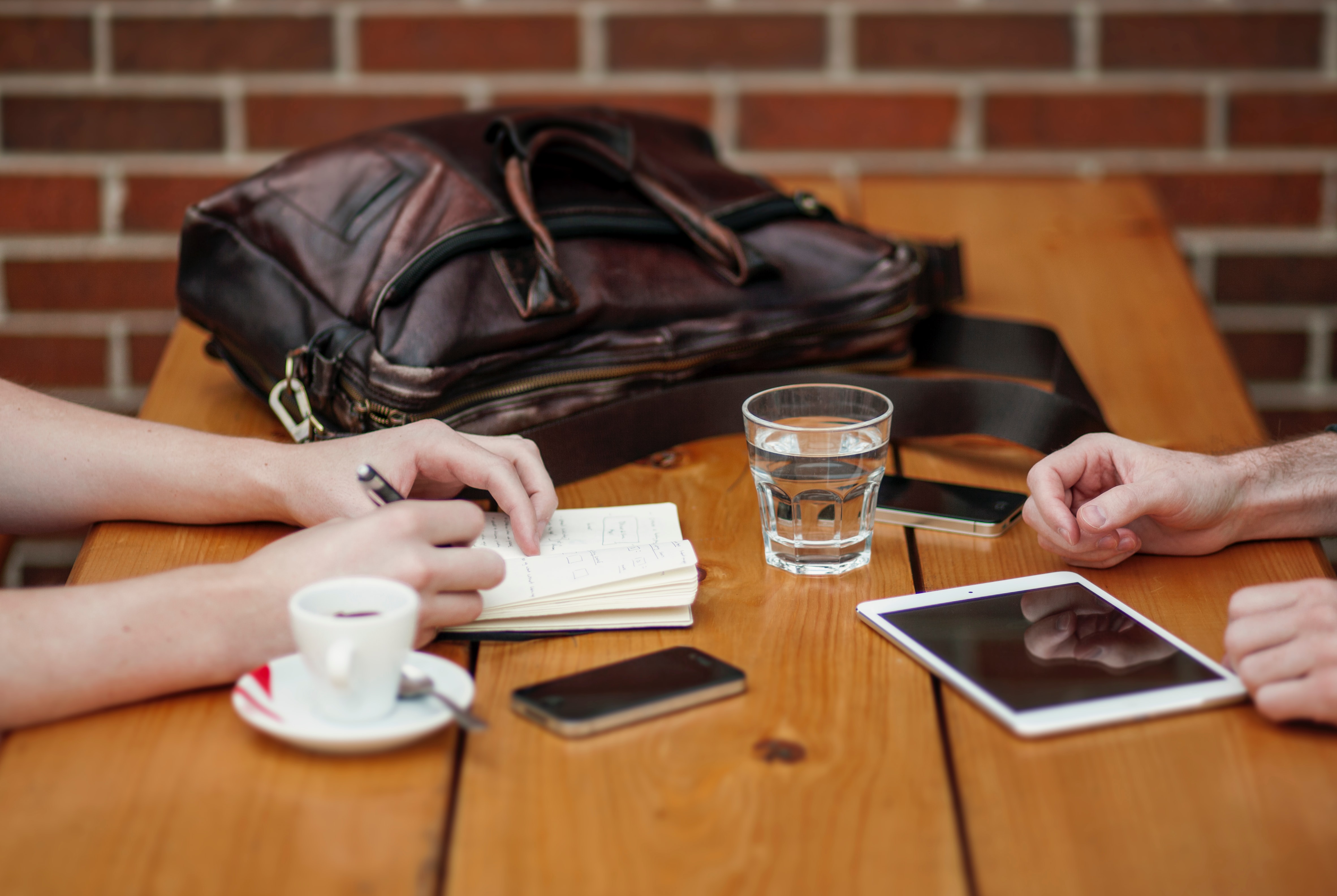 Two people are sitting across from each other with a large old leather-style satchel bag on the three large wooden plank table tops with a red brick wall at the end of the table. The person on the left is taking notes in a small notebook with an espresso coffee cup and a small iPhone to her right. The person on the righthas  two hands resting on the table top. There is a small glass of water and small iPhone in front of their right hand and an iPad mini in front of their left hand 